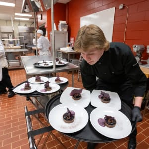 A young server in a black chef’s coat carefully arranges plated chocolate desserts with purple garnishes on a serving tray in a professional kitchen during the 2024 Celebrity Chefs Invitational. Other chefs in white coats work in the background
