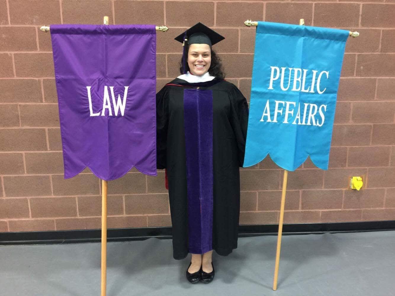 Melissa standing with two flags at graduation: Law (purple with white letterings) and Public Affairs (light blue with white letterings)