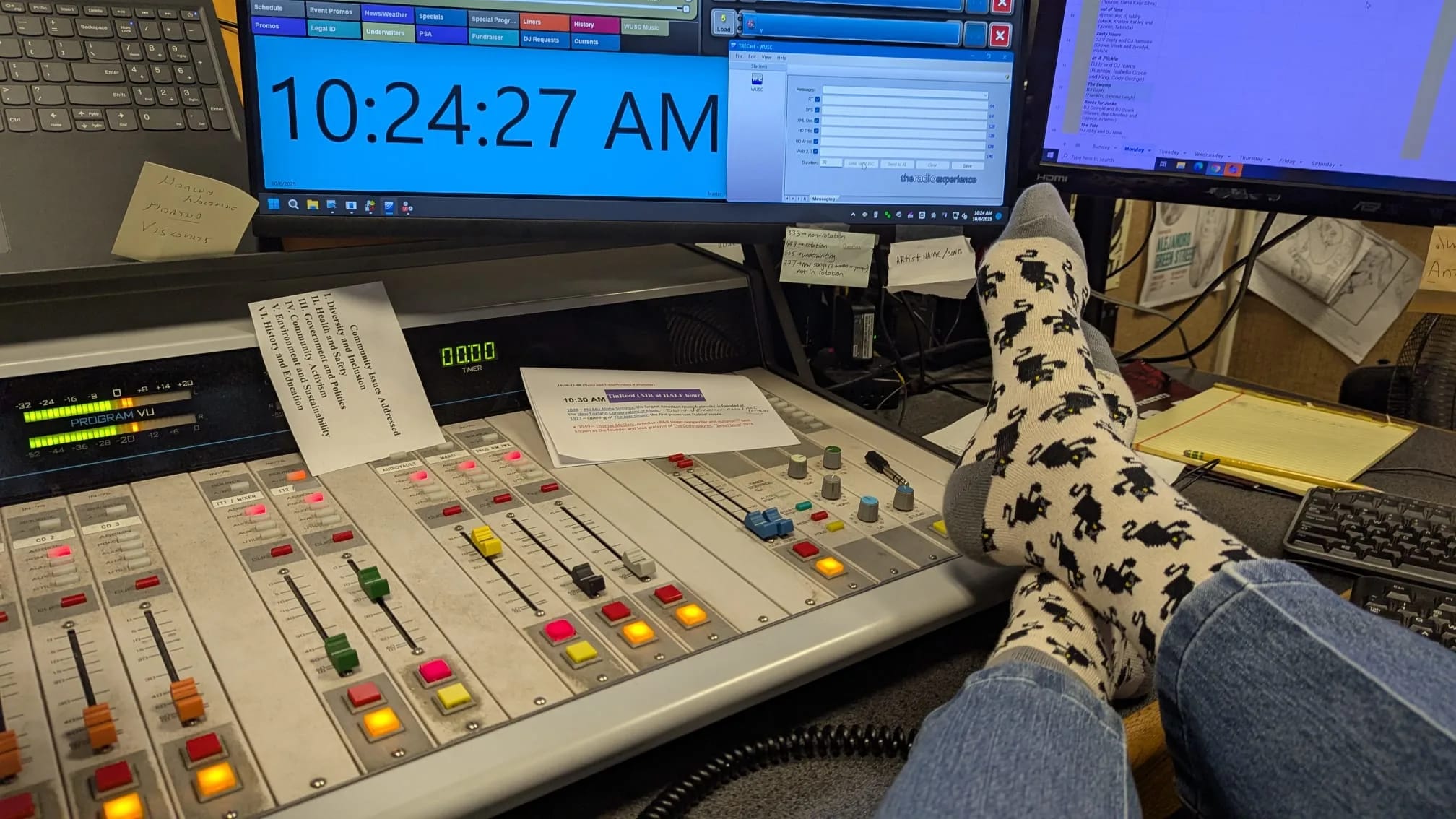 Feet in patterned socks rest on a radio studio mixing board; computer monitors display time and audio software.
