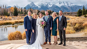 A pair of newlyweds and guests on a clearing before a large lake and snowy mountain range