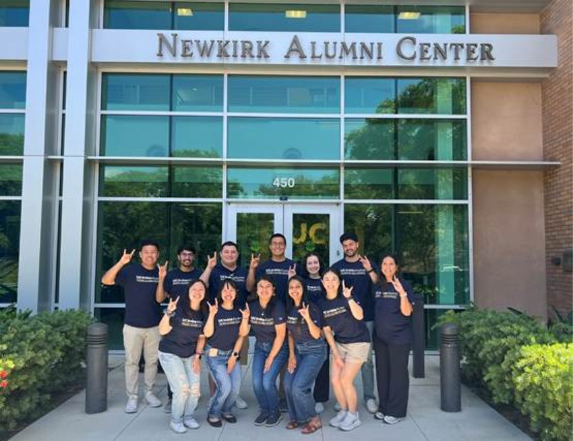 2025-26 Young Alumni Council smiling and holding up zots in matching navy blue shirts in front of the Newkirk Alumni Center.