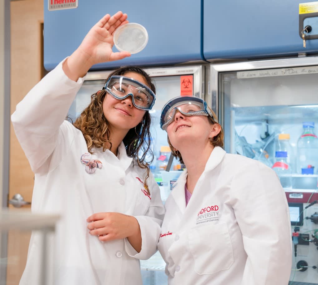 Two students with goggles looking up at a petri dish