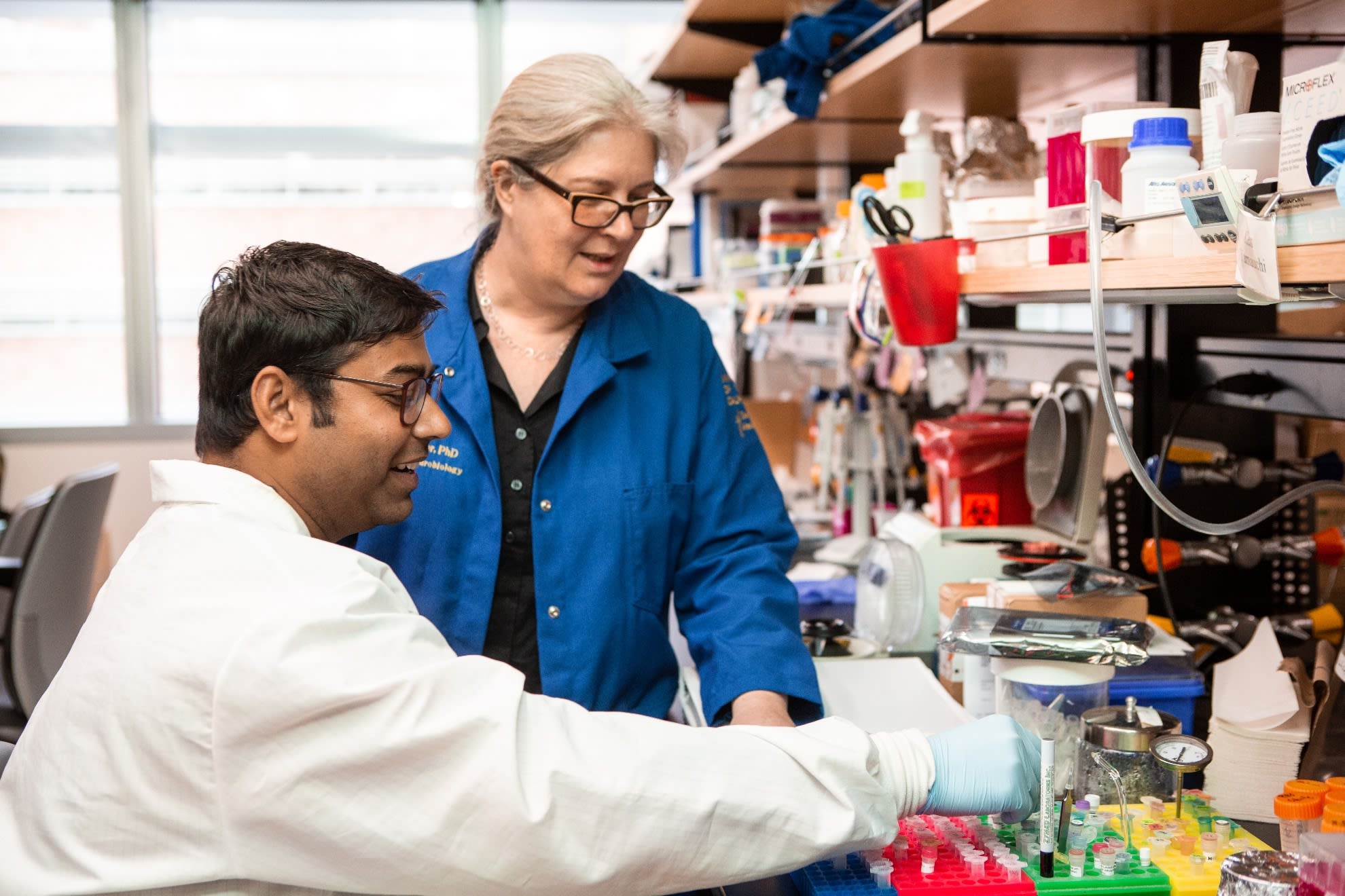 One standing researcher oversees another researcher as he sits and picks up a small plastic tube.