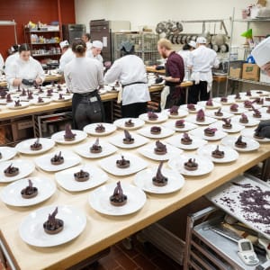 Culinary students in white chef coats and hats prepare elegant plated desserts in a professional kitchen at the 2024 Celebrity Chefs Invitational. Rows of meticulously plated chocolate tarts with purple garnishes line the tables as students focus on final touches.