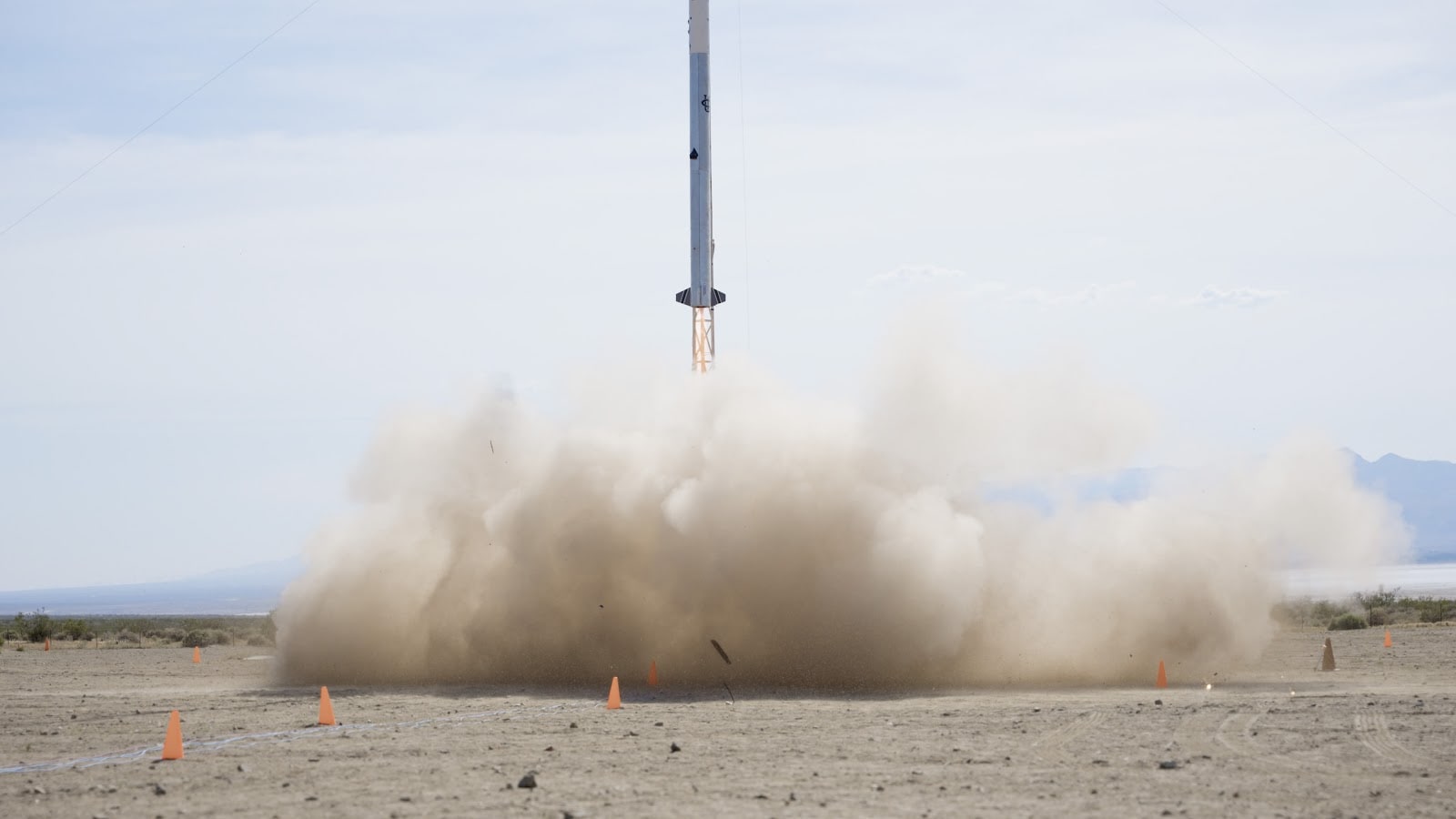Image of PTR Launch, 2023: showing the bottom half of the rocket as it launches; there is a cloud of dust and several small orange cones over the launch sight. The landscape beyond has the look of a desert in the middle of the day.