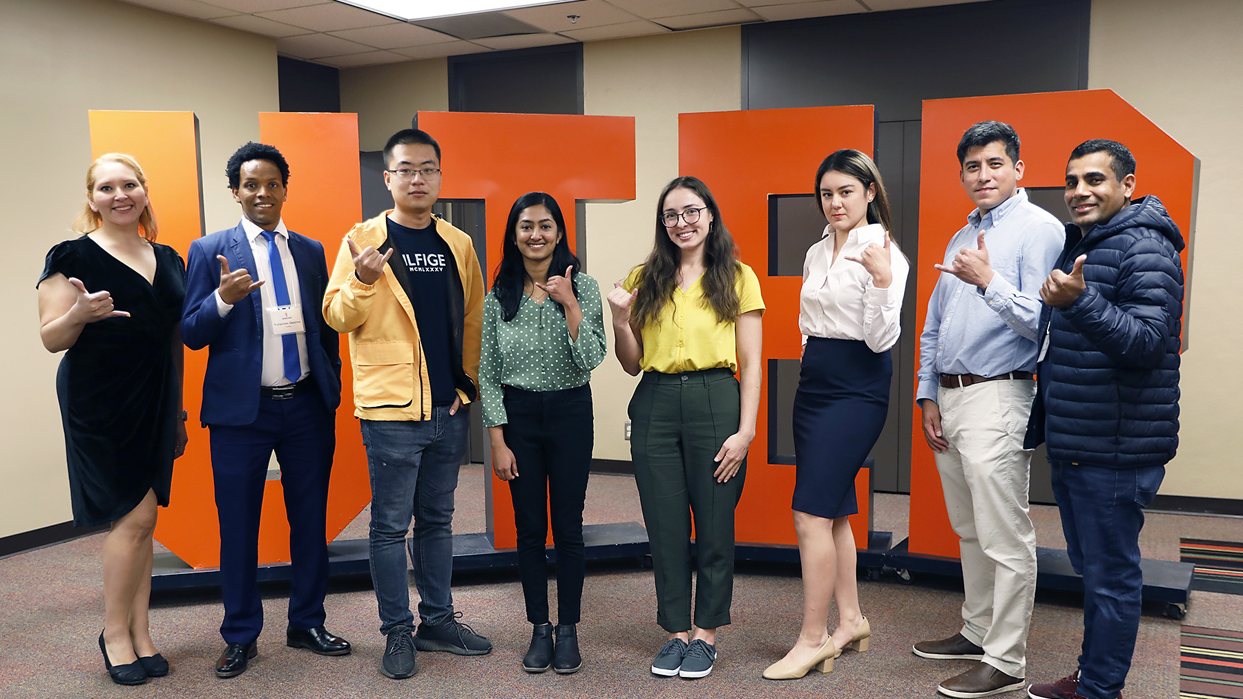 Eight graduate students at Minute Thesis event, posing with picks up in front of giant UTEP sign