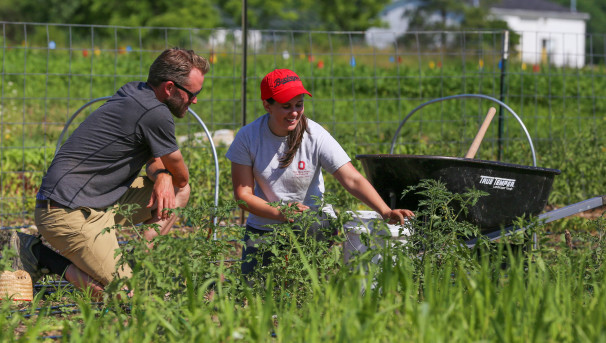 Two individuals kneeled in the CFAES Student Farm. One holds onto the edge of a large, white bucket