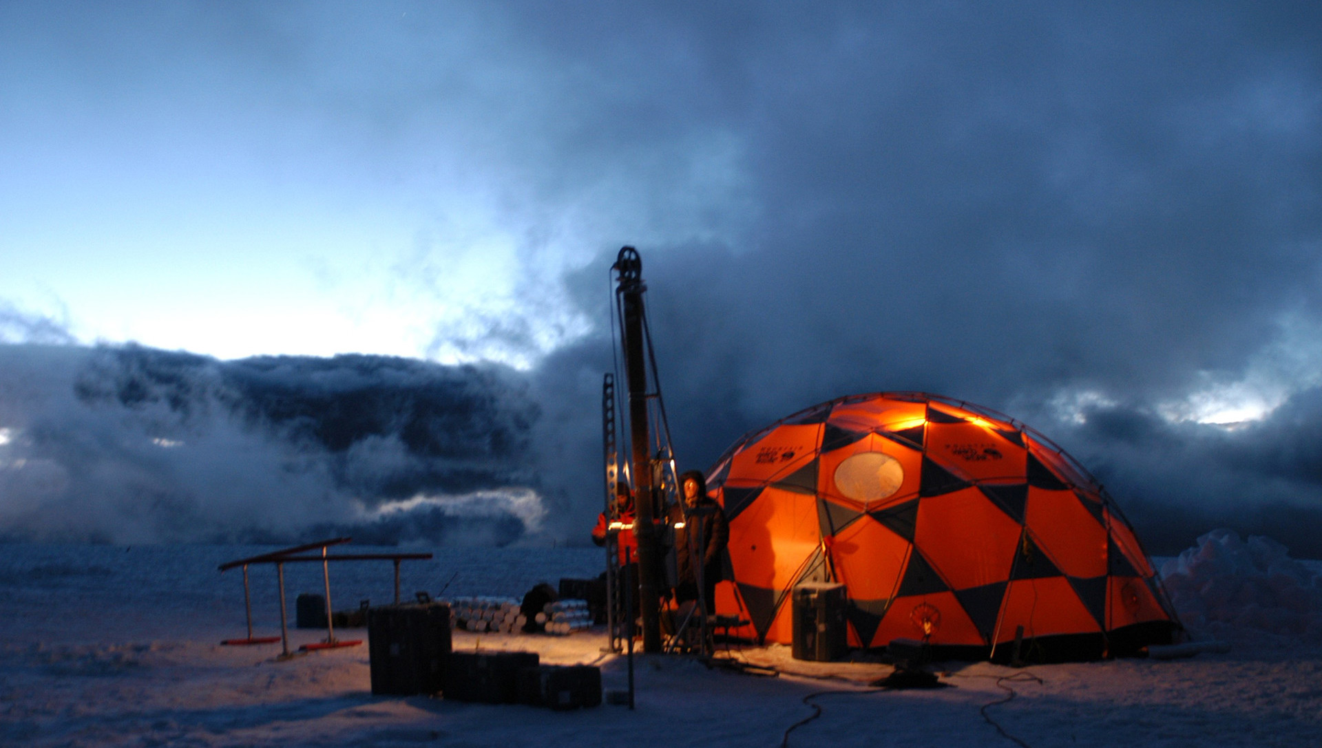 Two researchers stands outside their tent, the sun setting on the snowy environment