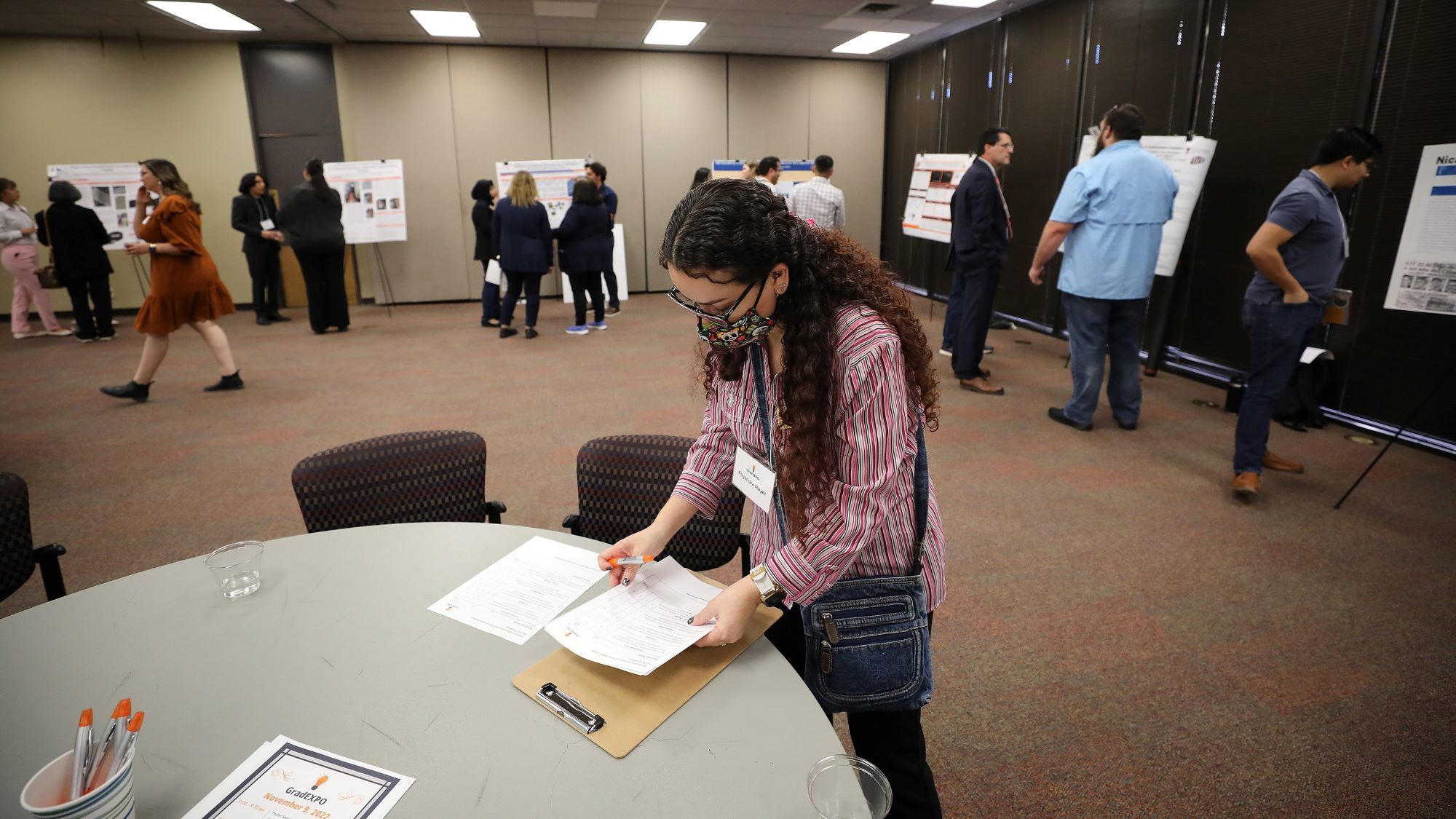 Woman filling out paperwork at Grad Expo table, surrounded by people viewing academic posters