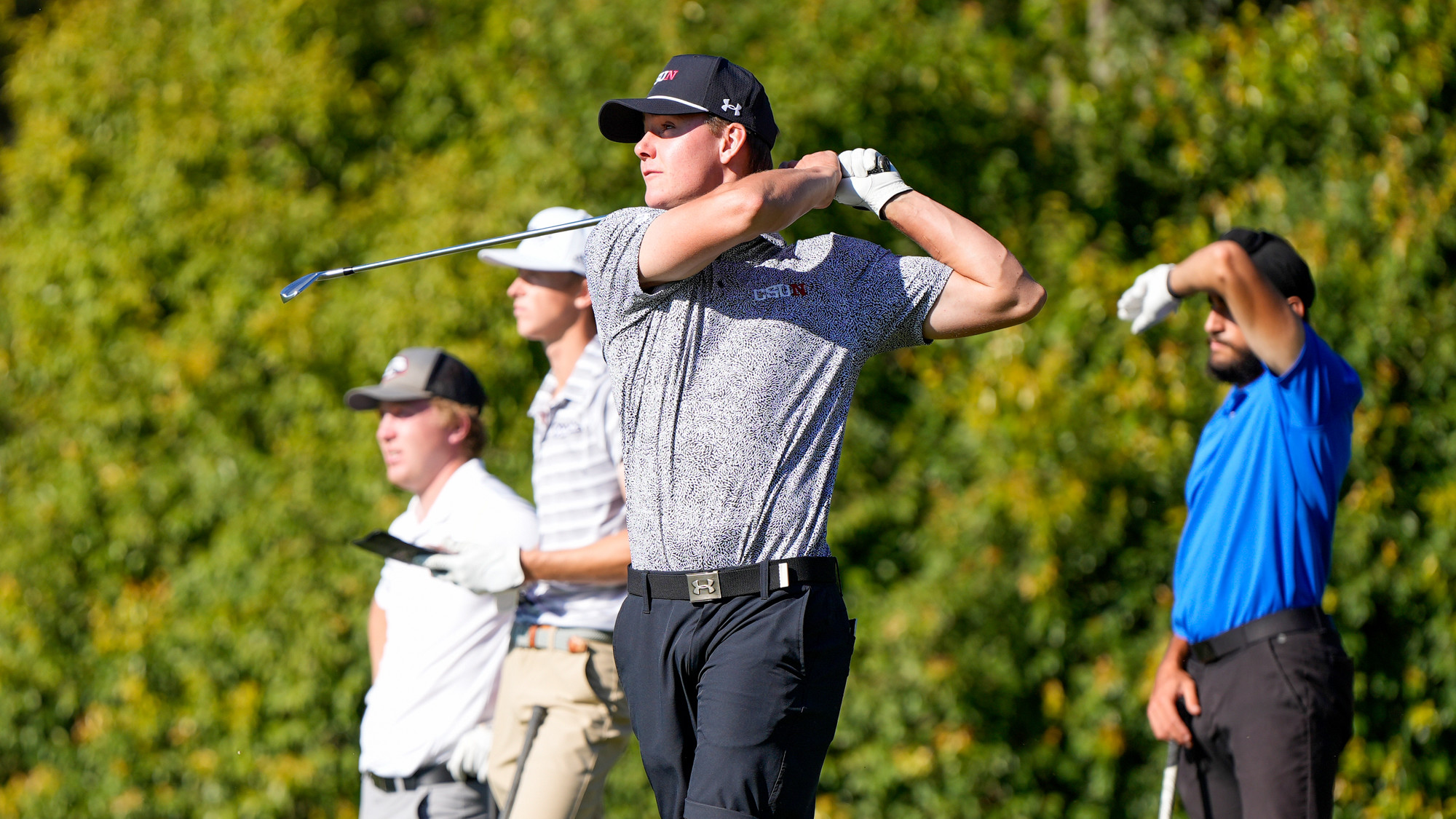 A golfer stands on the course holding a putter with trees and greenery in the background.