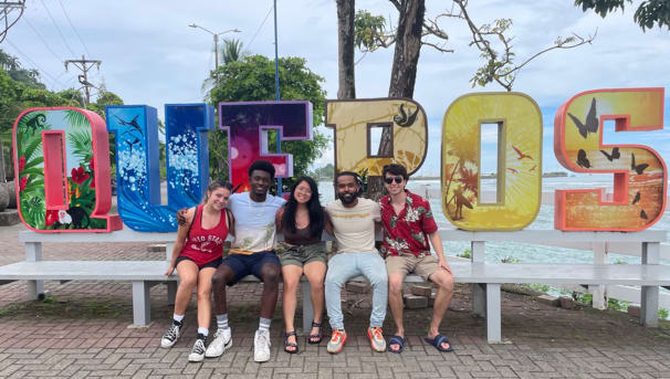 Five students sitting on a bench in front of a sign.