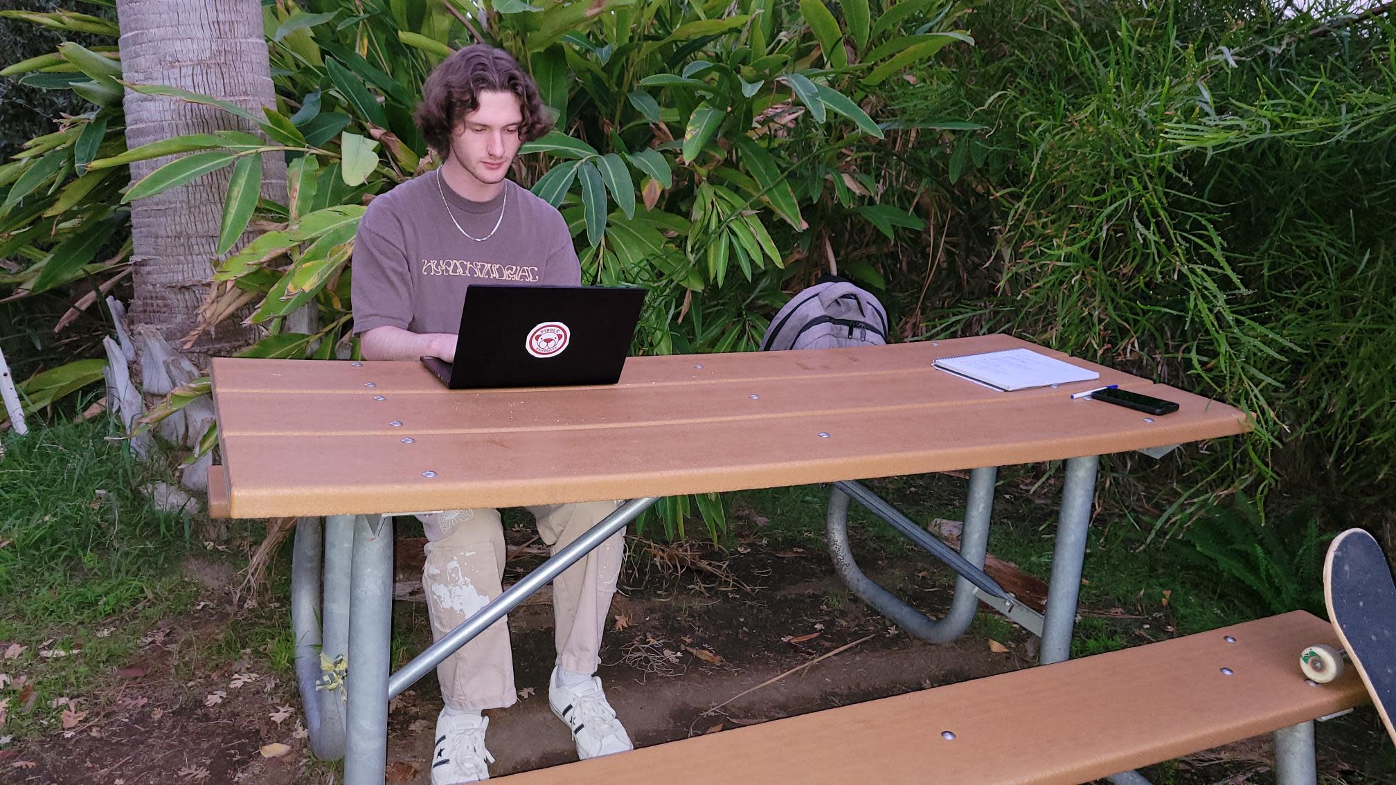 Student works on a laptop at an outdoor table surrounded by greenery.
