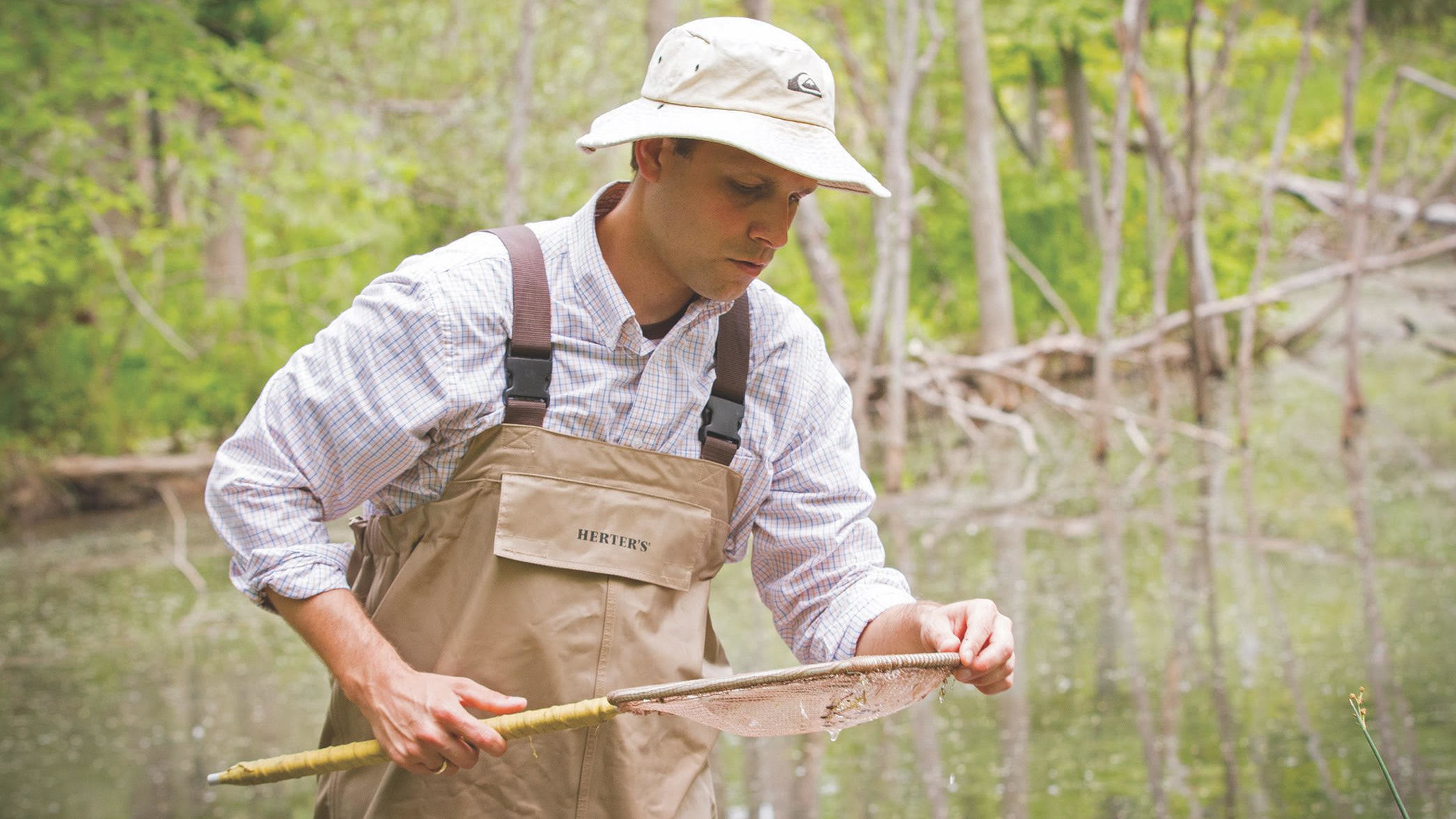 An OU student wearing tan waders stands in marshy water while inspecting a net.