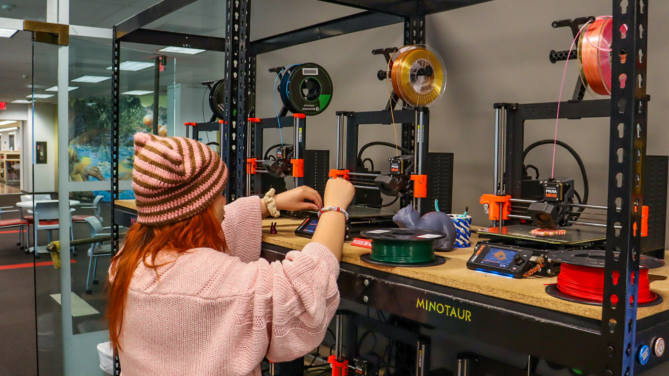 Student in pink sweater works on a 3D printer, one of several lined up on a shelf.