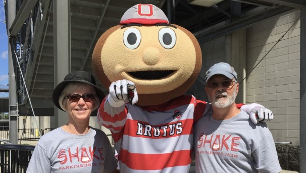 Two individuals stand beside Brutus Buckeye, who points towards the camera
