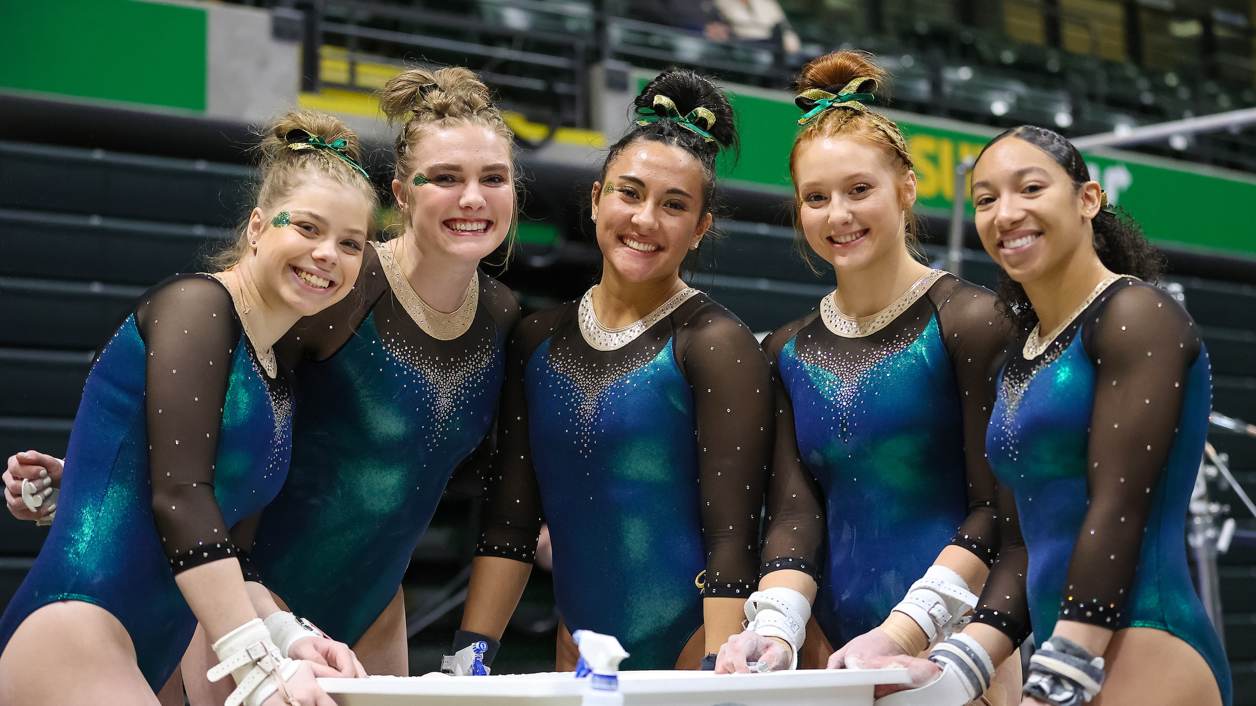 UAA Gymnastics team, 5 teammates stand together in shiny blue and green uniforms