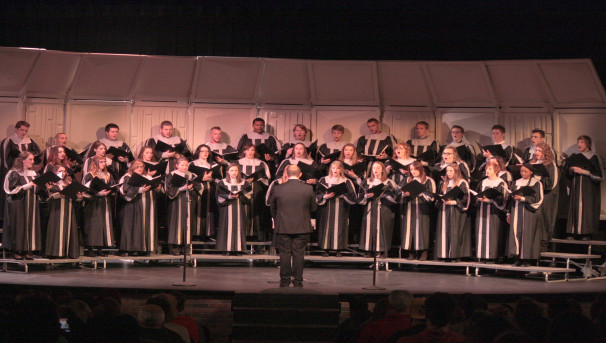 A large group of Lima Chorale students sing on stage, holding black folders