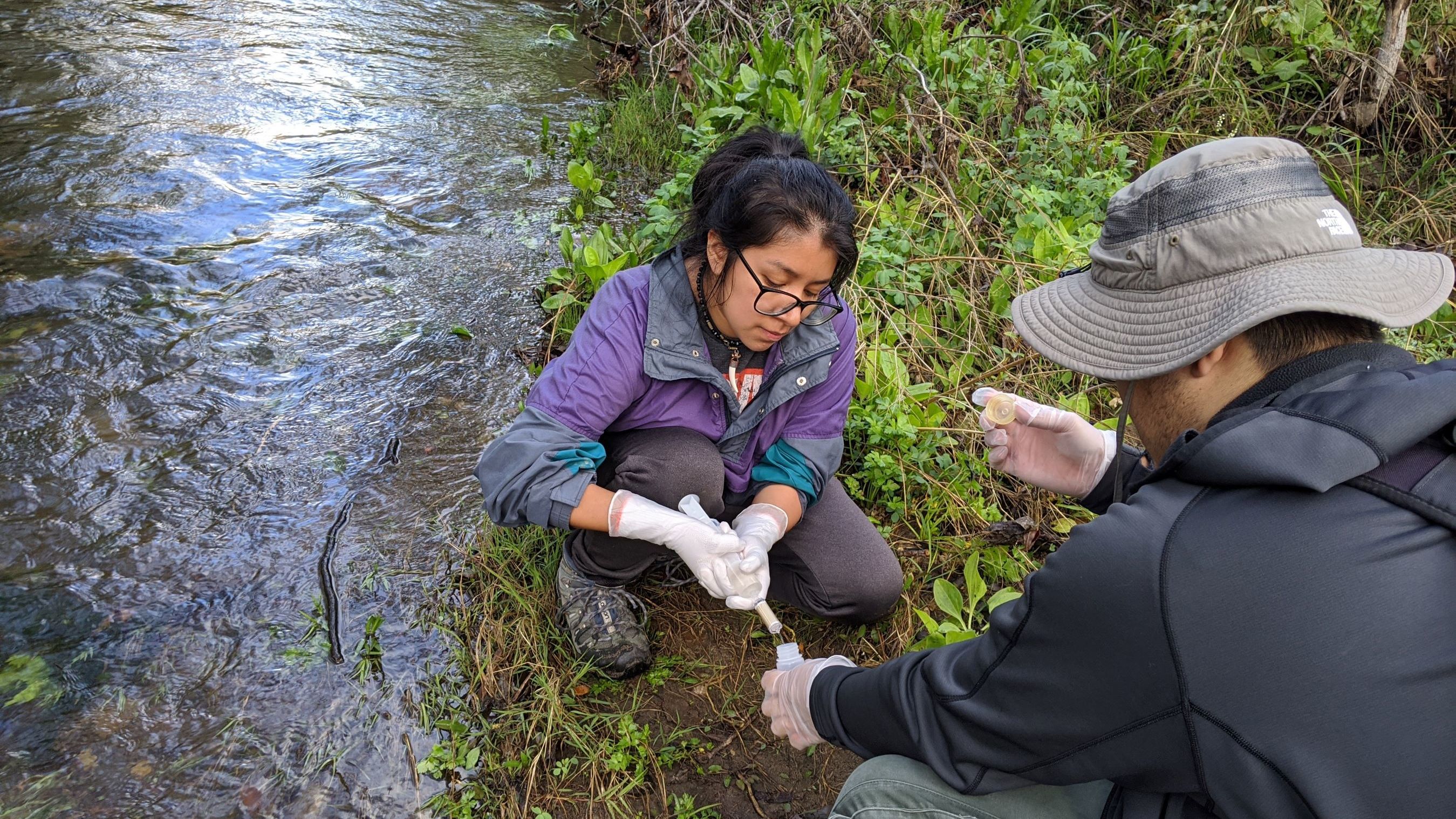 Two researchers in lab coats prepare sample bottles together at a workstation.