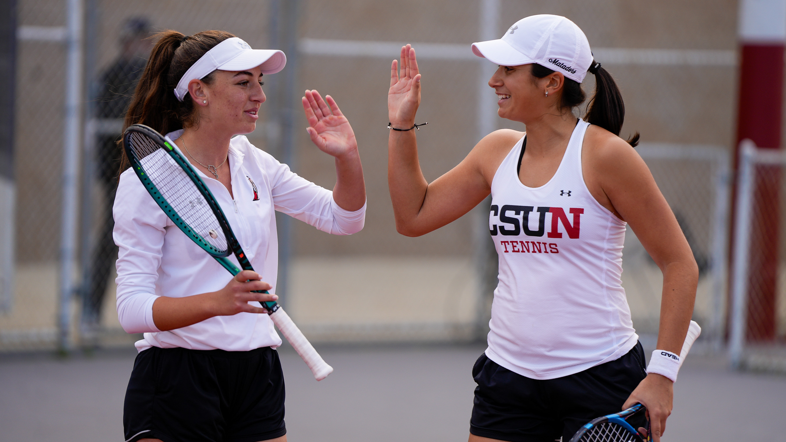 Two tennis players high‑five on the court, holding rackets and wearing visors.