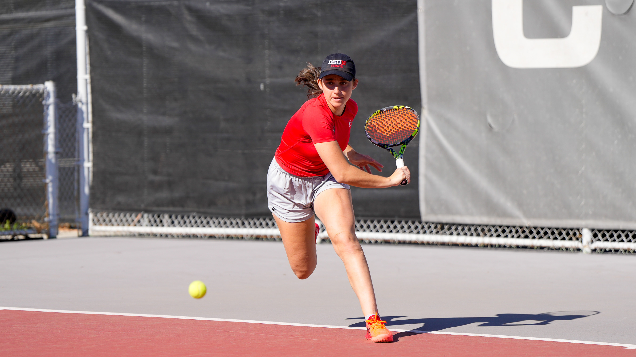 A tennis player in a red shirt and gray shorts stretches forward to return a ball.