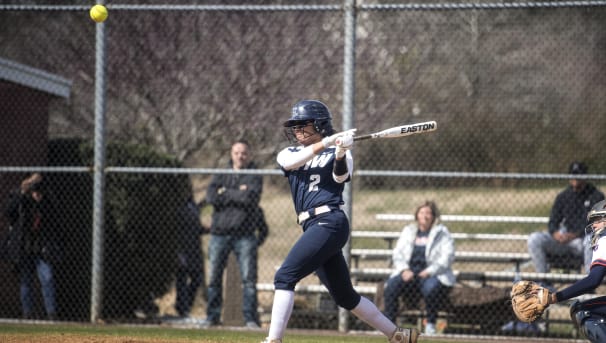 A softball player is getting ready to take a swing as the ball flies through the air.