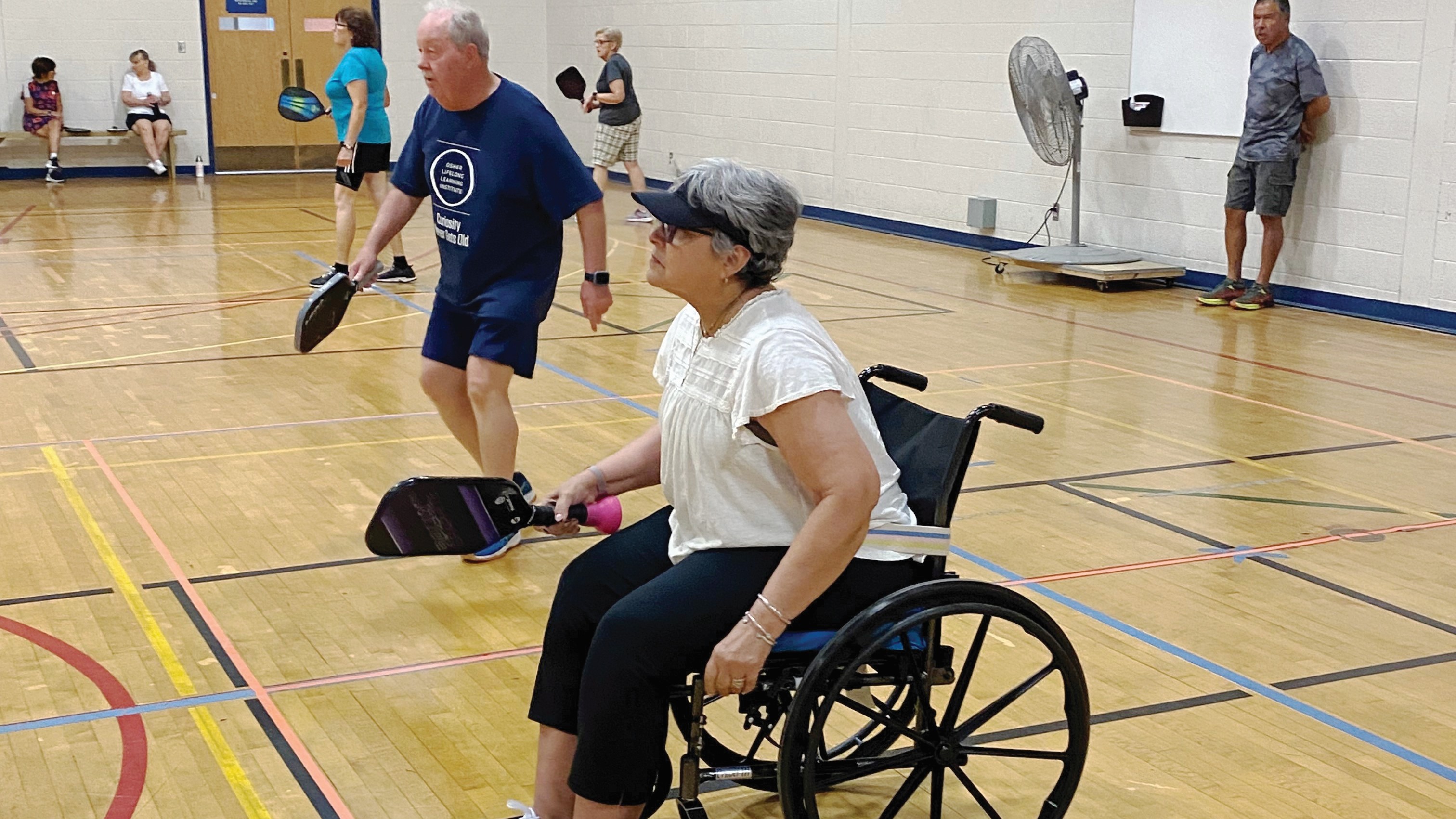 Woman in a wheel chair and man standing, ready to play pickle ball with their paddles.