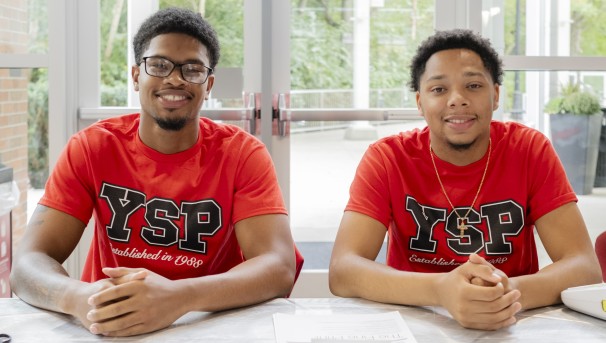 Two students sitting at a table with their hands clasped, wearing Young Scholars Program t-shirts.