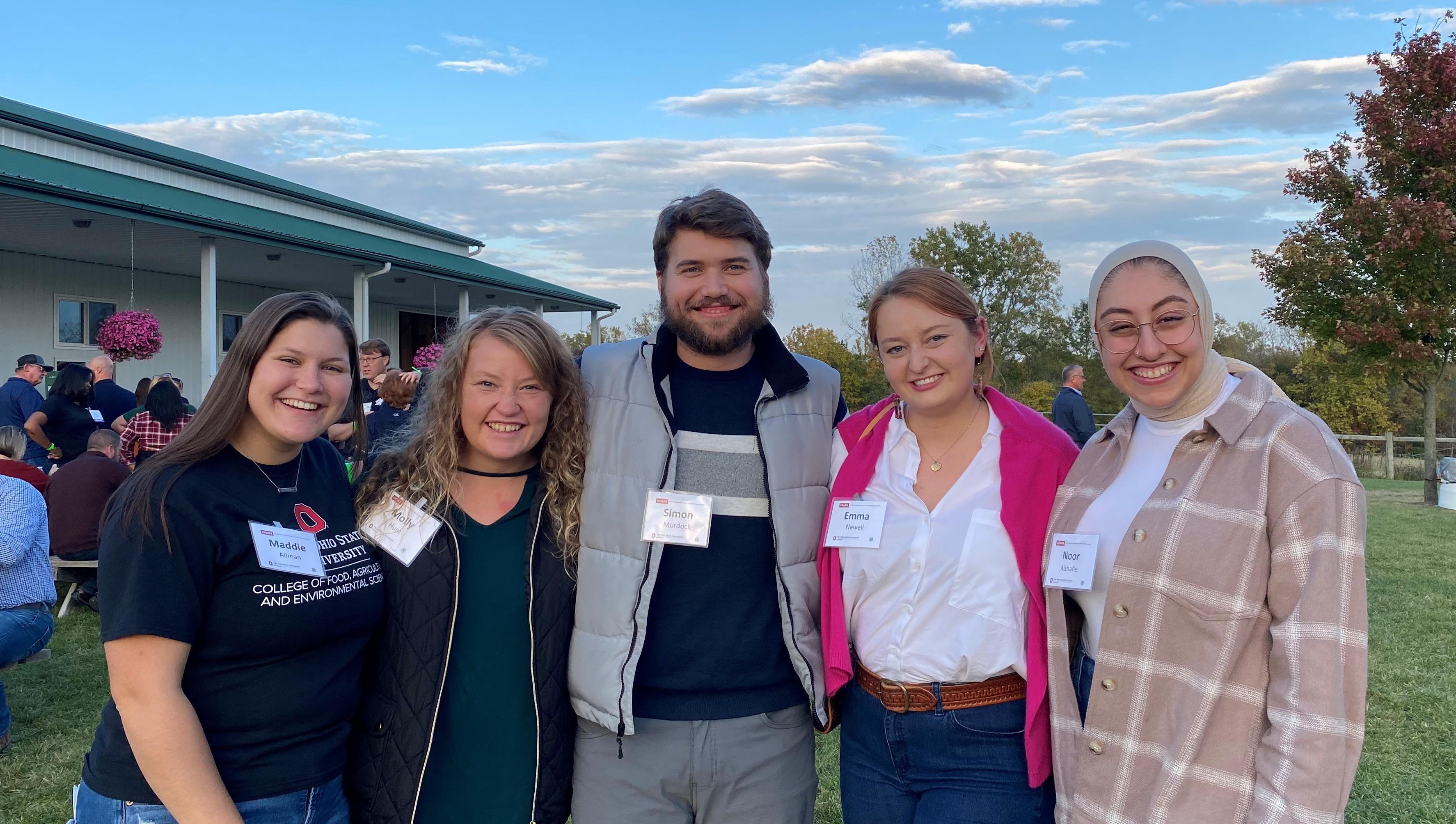 Five young adults are standing outside on a farm smiling together.