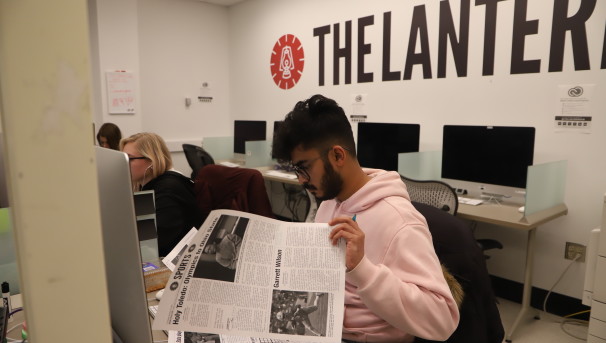 A student sits before a large desktop computer. He is flipping through The Lantern's newspaper