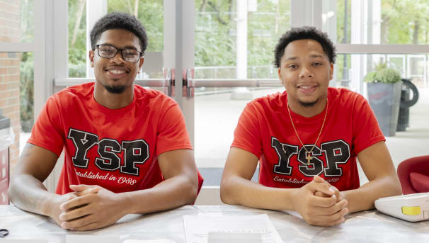 Two students sitting at a table with their hands clasped, wearing Young Scholars Program t-shirts.