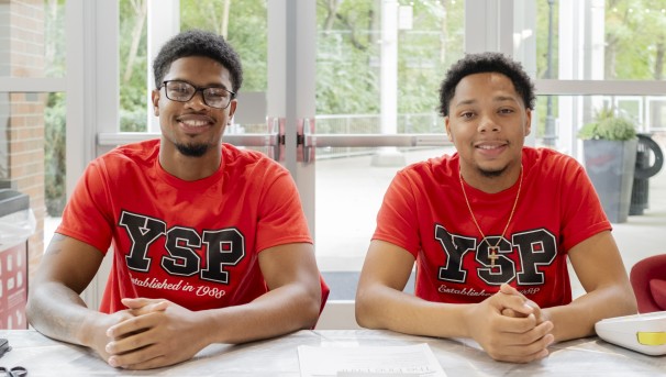 Two students sitting at a table with their hands clasped, wearing Young Scholars Program t-shirts.