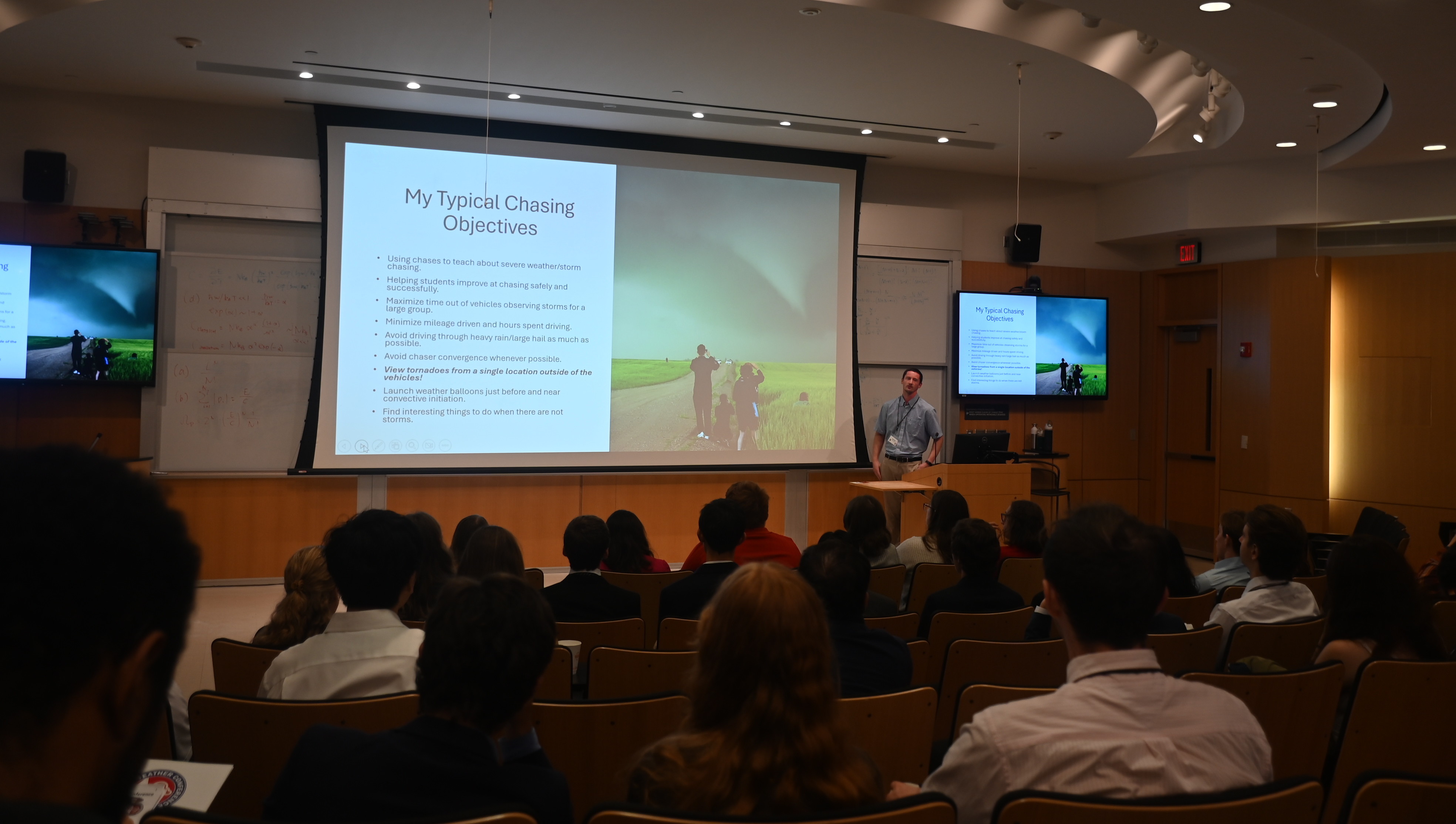 A man stands at a podium, presenting to an audience, with a large screen showing a PowerPoint slide.