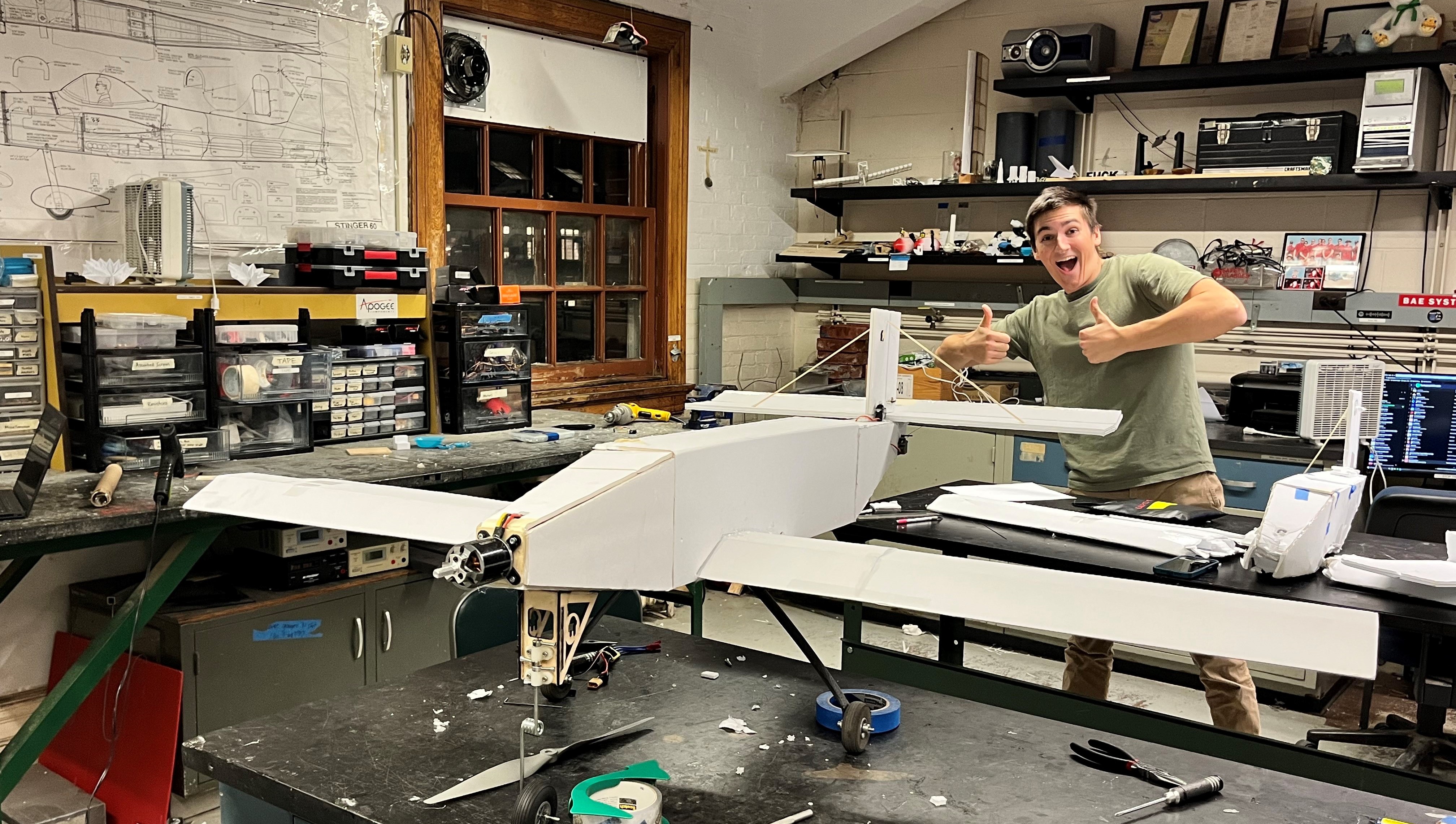 A student giving two thumbs up in a workshop with a model plane.