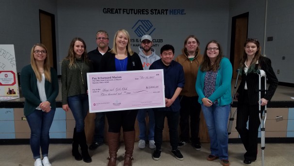 Nine individuals standing around a large, pink check for Pay It Forward Marion