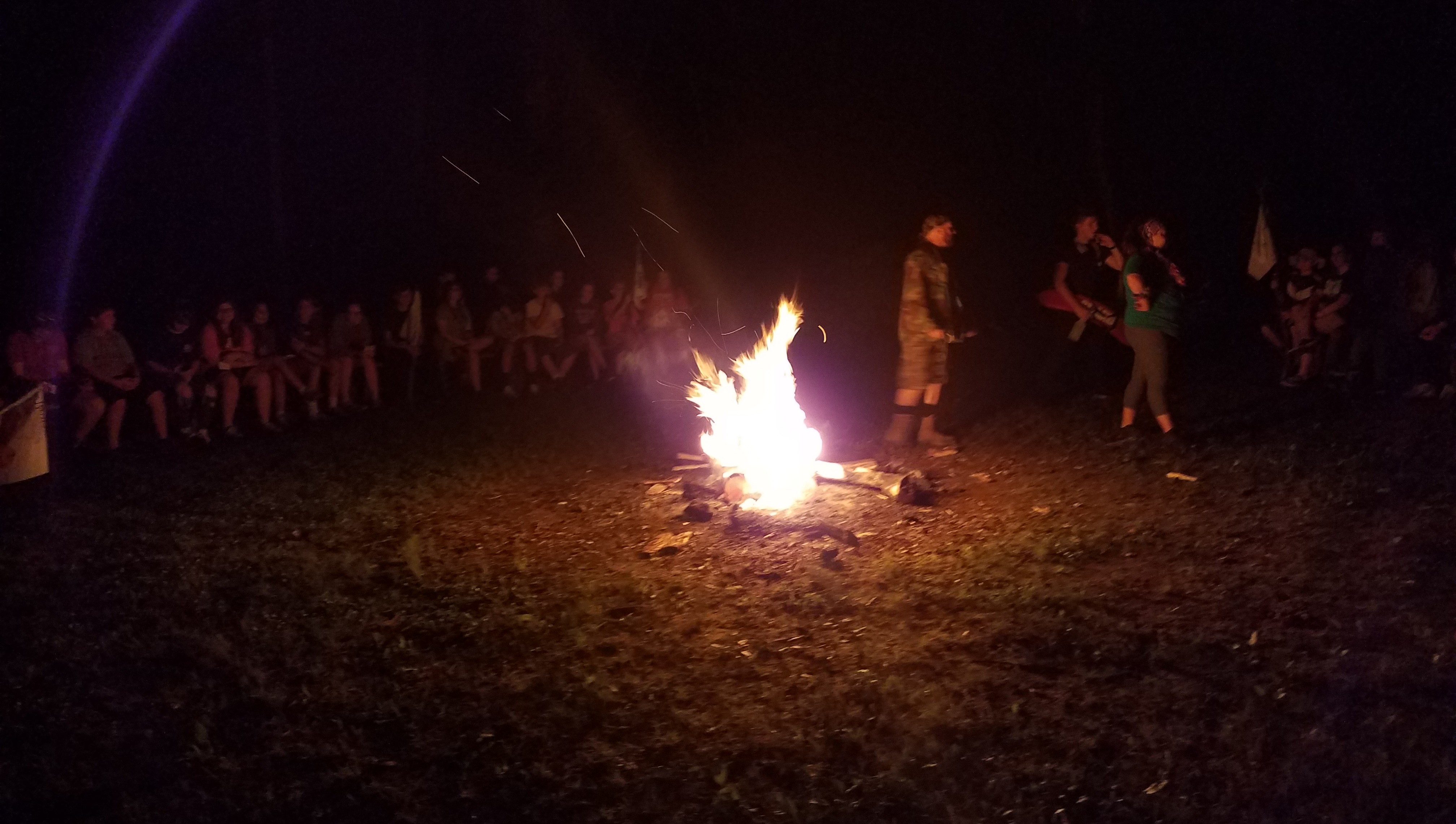 Campers and staff sit outside at night, surrounding a large, lit firepit