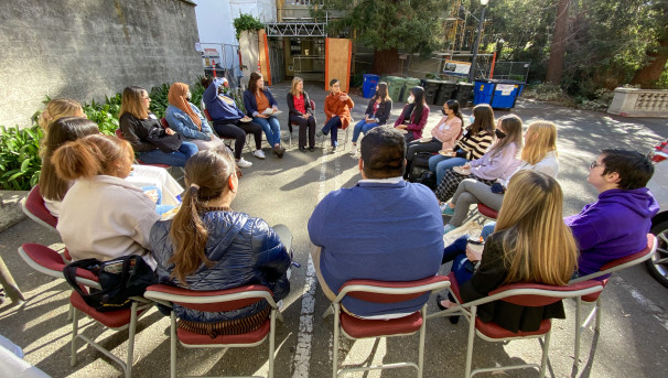 20 people sit in a circle outside on a sunny day.