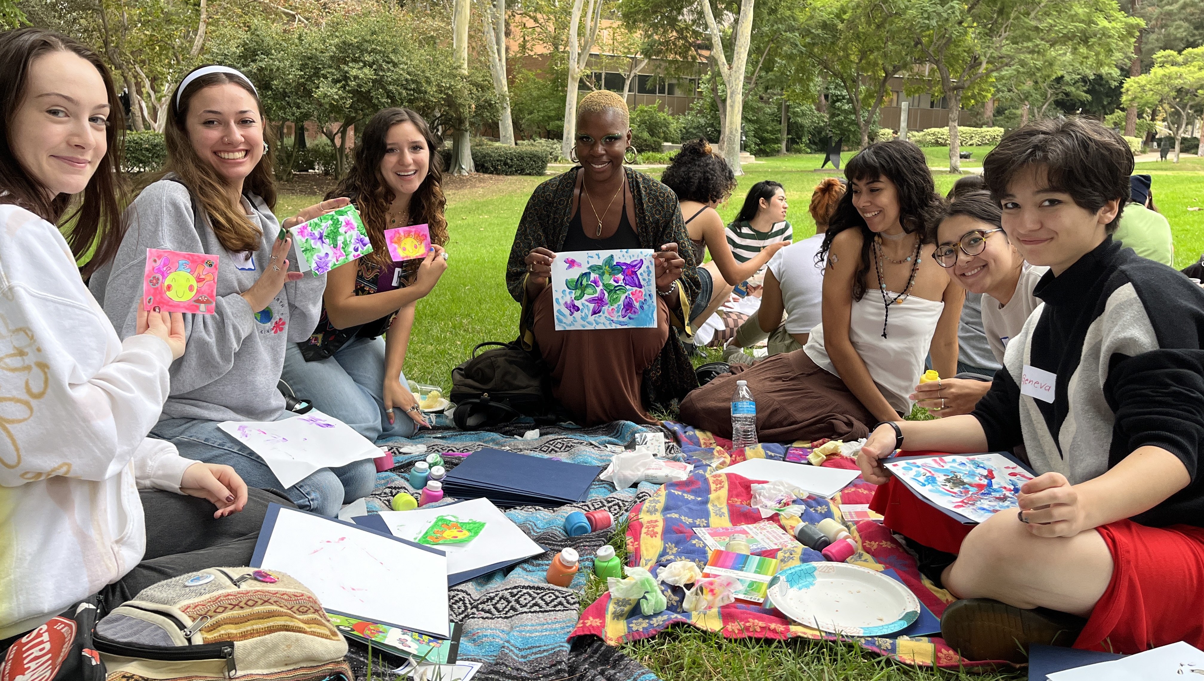 a group of people sitting on the grass, smiling, and showing off their paintings