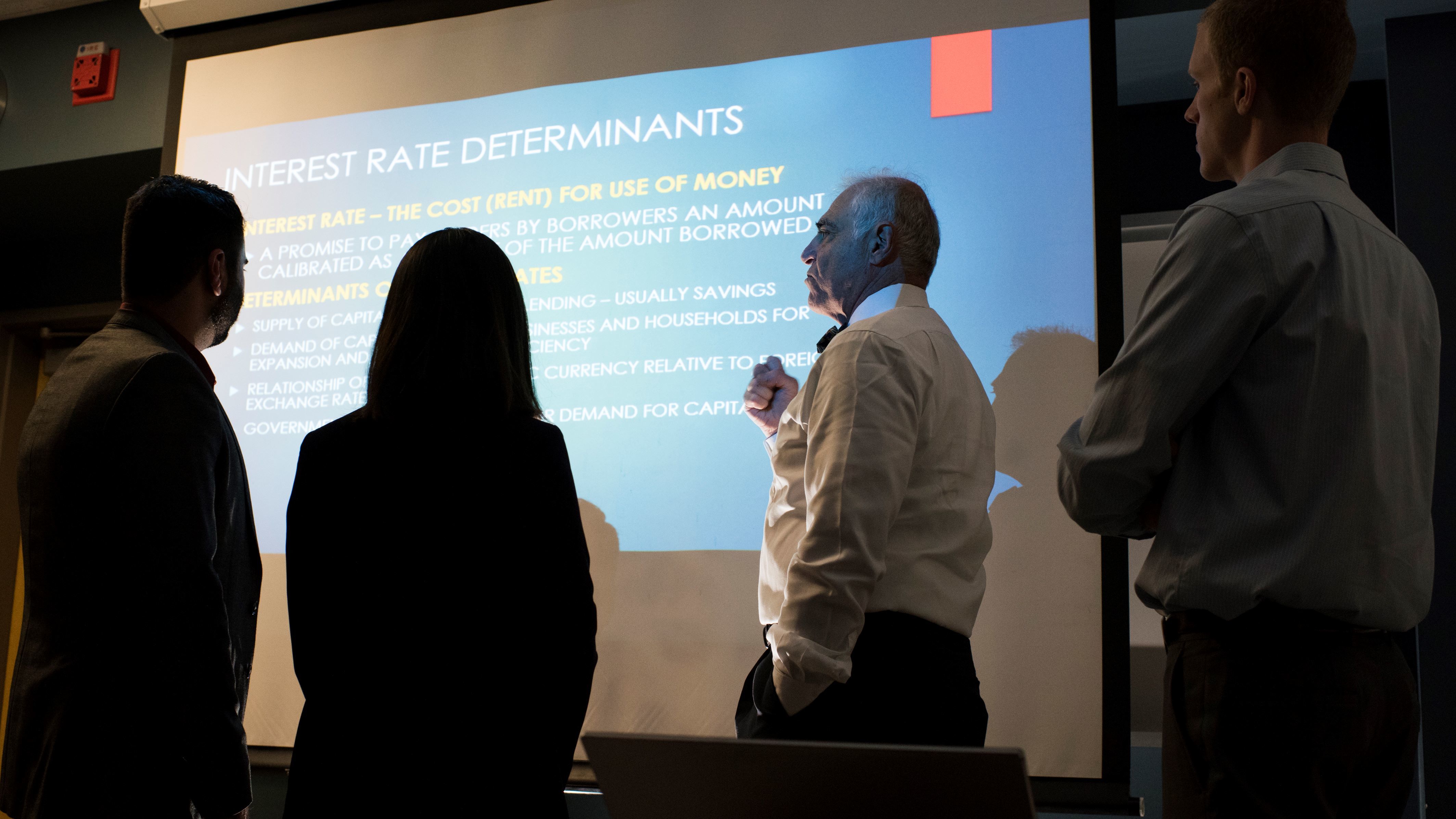 Professor lectures on interest rate determinants as students observe a projected presentation.