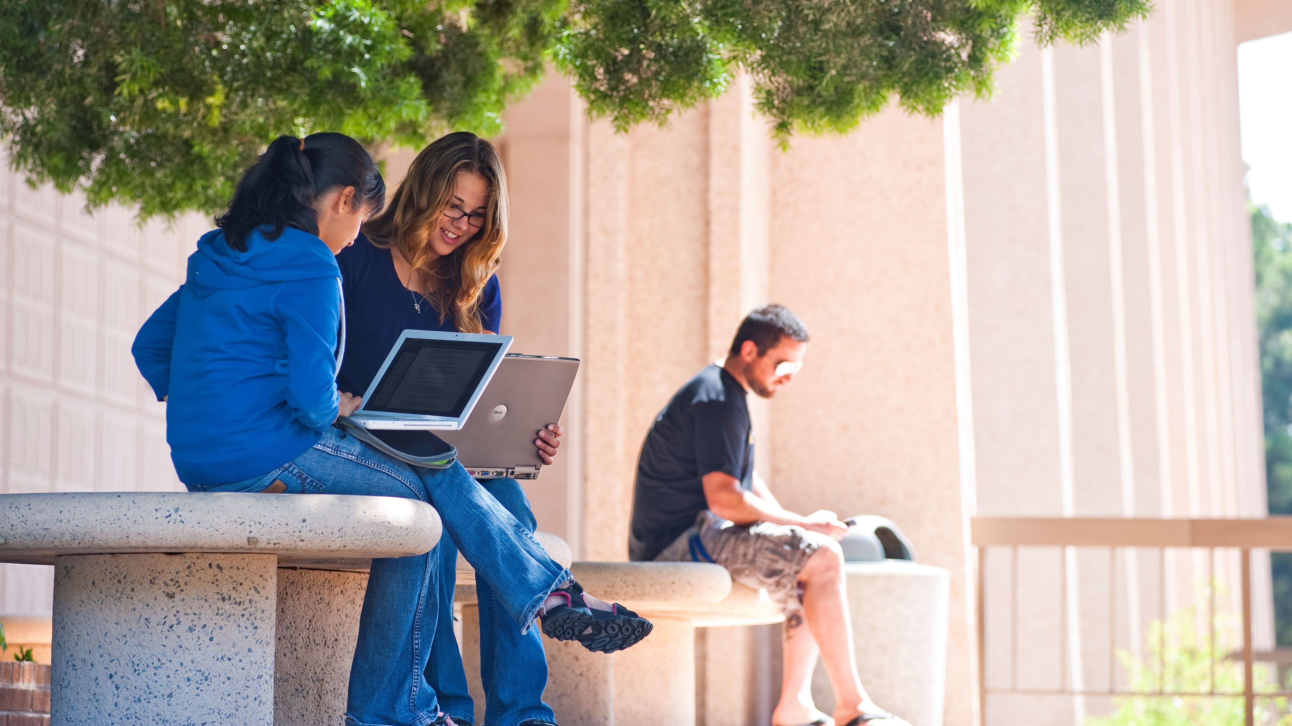 Students sitting in front of the University Library on a bench, working on a laptop