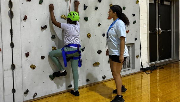 Camper on a climbing wall wiearing helmet and belaying gear.