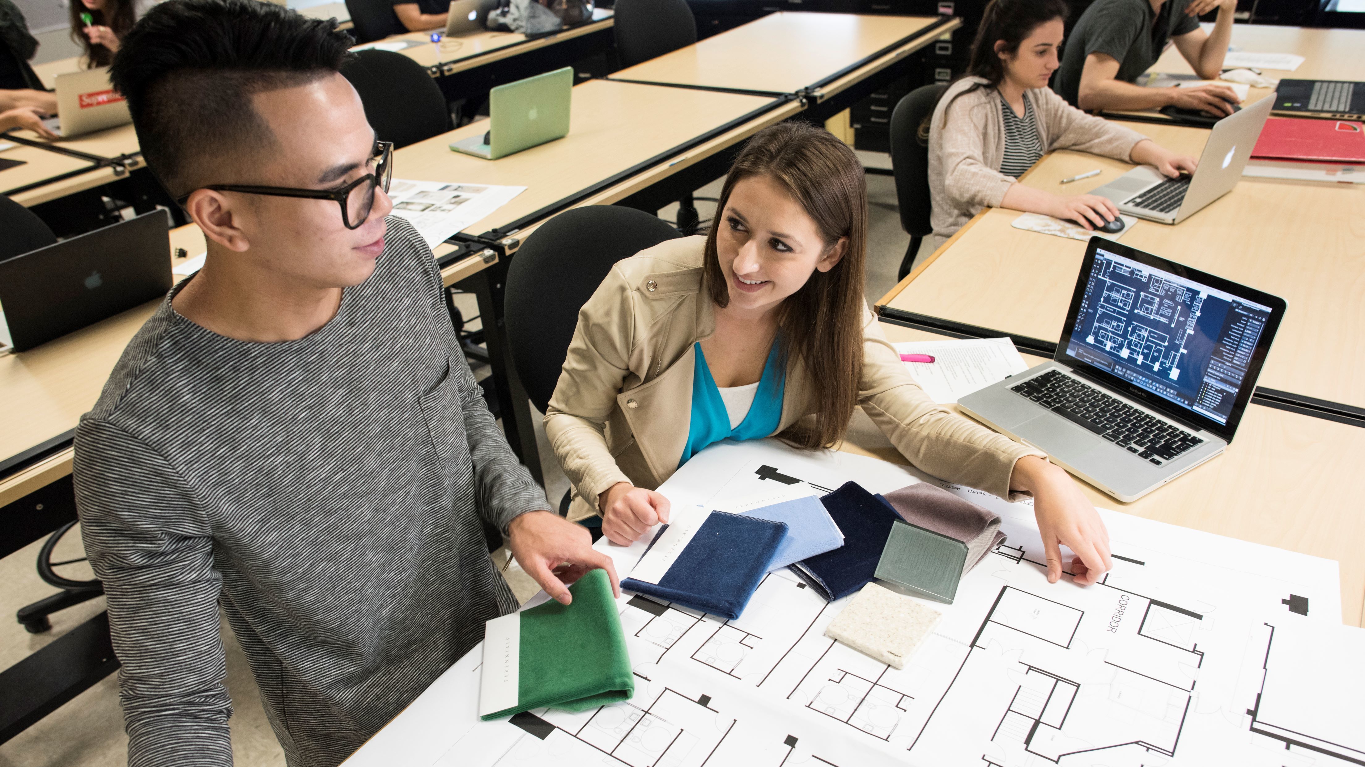 Two students working on a laptop and architectural designs.