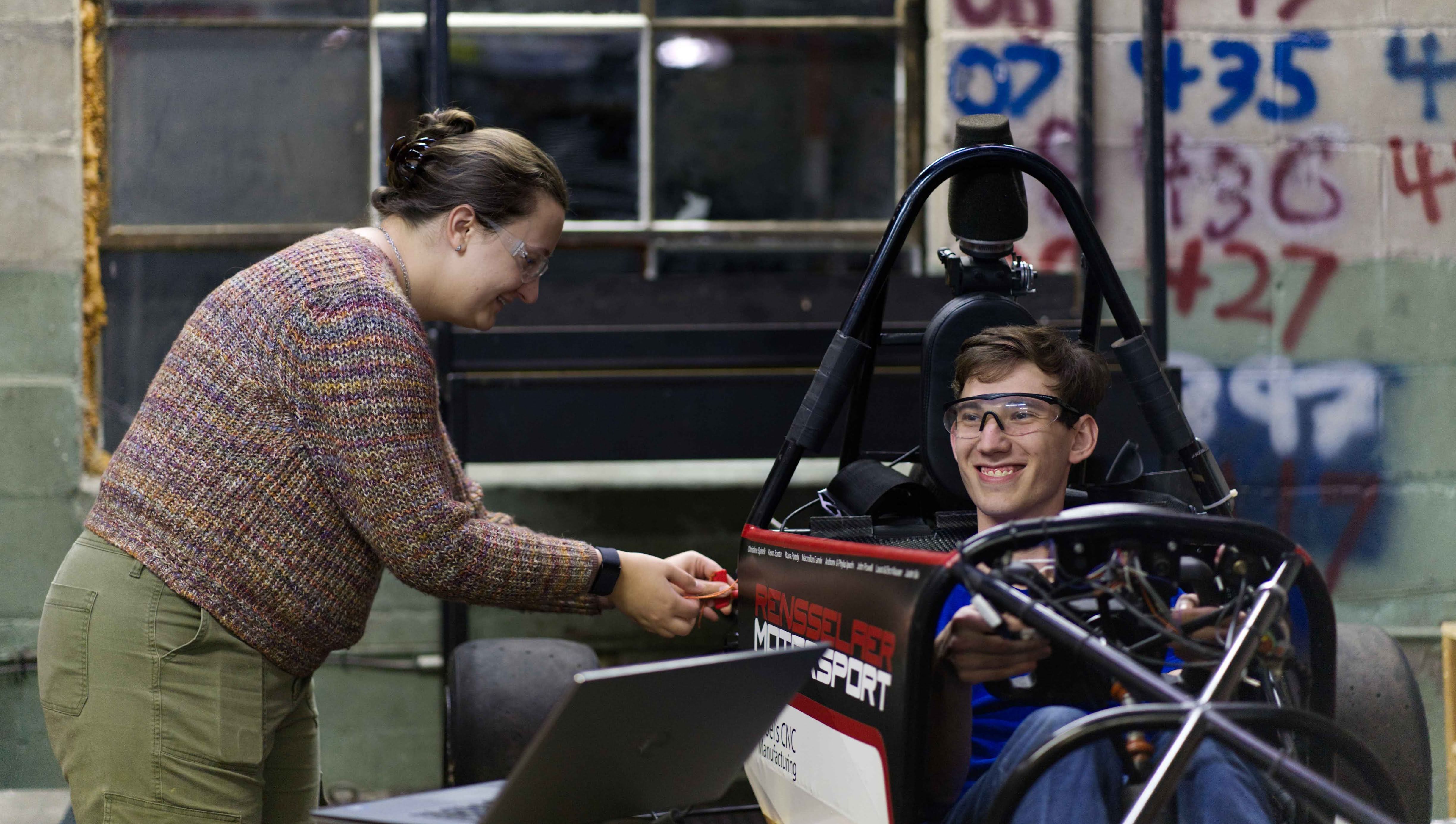 A student smiling while sitting in a former Motorsport vehicle, with someone working on the side.