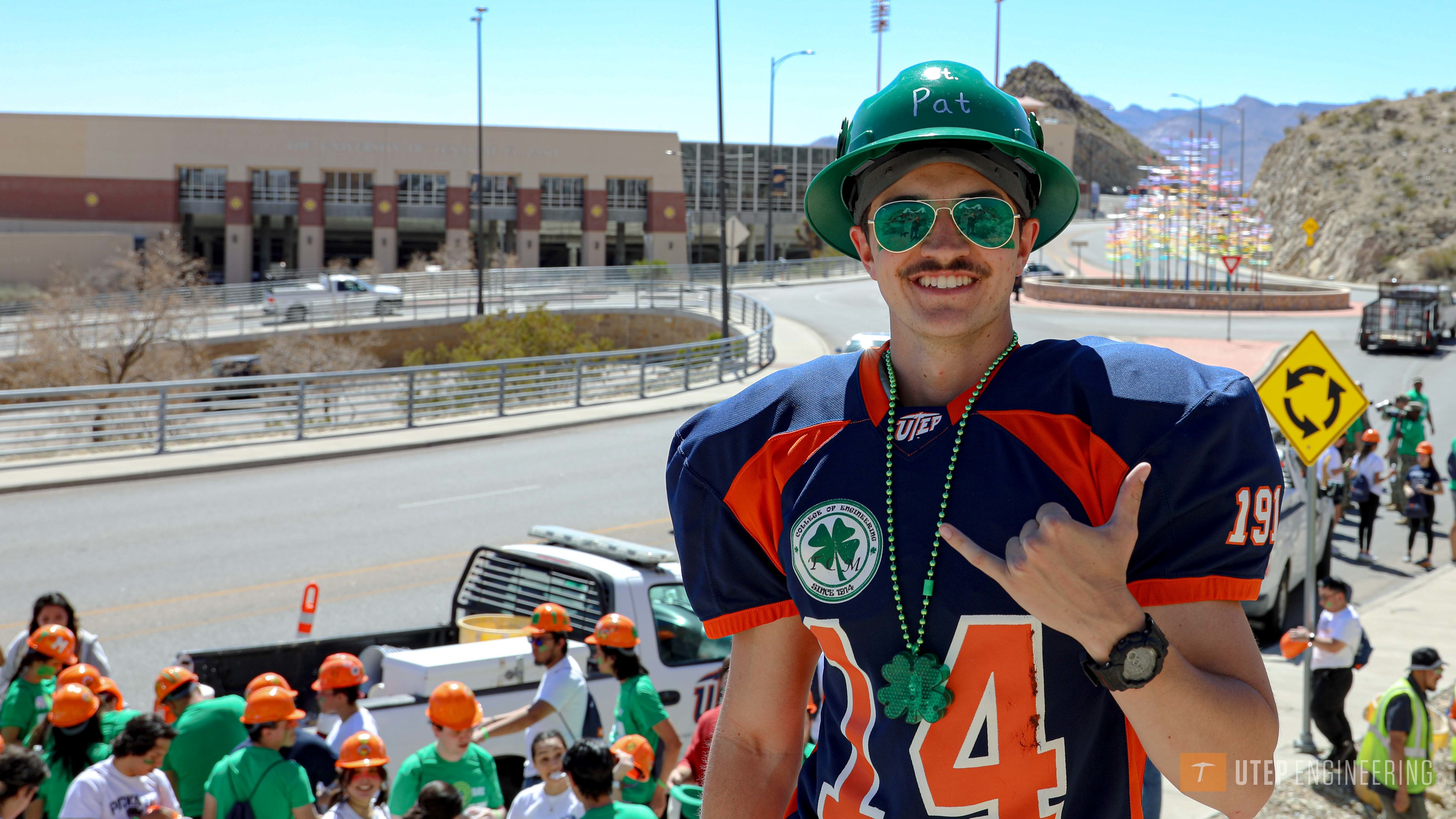 Male student with mustache, green hard hat labelled Pat, green sunglasses, in UTEP football jersey