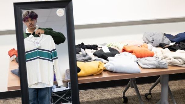 Student holding up a shirt in a mirror at the gender affirming wardrobe.