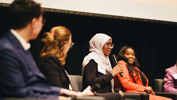 Student talking into a microphone to an audience while other students on panel actively listen