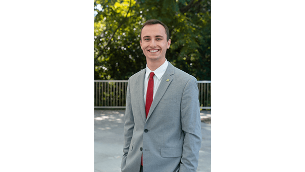 Max Wilberding's professional headshot. He wears a grey suit with a deep red tie