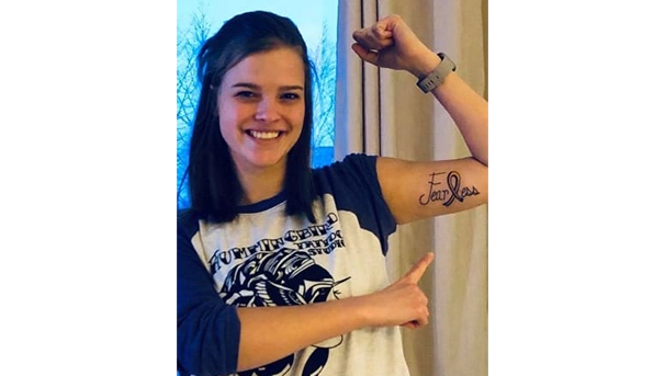 A woman points to a tattoo on her inner arm, which says 'Fearless.' The L is a cancer ribbon