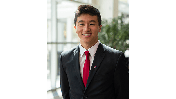 Patrick Lee's professional headshot. He wears a black blazer, a white button up, and a red tie