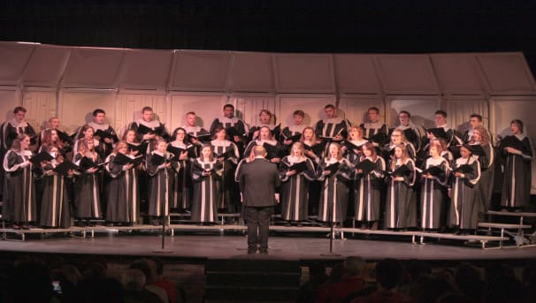 A large choir group sings on stage. They hold binders of music and wear black and white robes