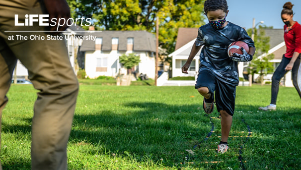 A masked boy holds a football while working on foot movement. He is in a grassy opening